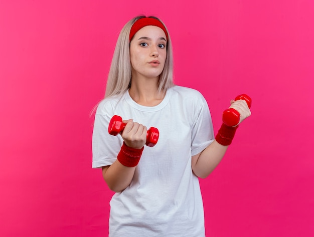 Impressionné jeune femme sportive avec des accolades portant un bandeau et des bracelets détient des haltères isolés sur un mur rose