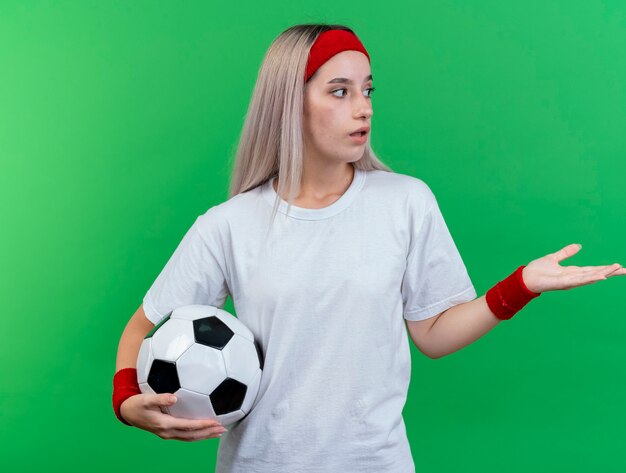 Impressionné jeune femme sportive avec des accolades portant bandeau et bracelets tient la balle à la recherche et pointant sur le côté isolé sur mur vert