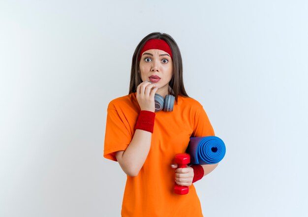 Impressionné jeune femme sportive portant bandeau et bracelets et écouteurs sur le cou tenant haltère avec tapis de yoga touchant le menton à la recherche d'isolement