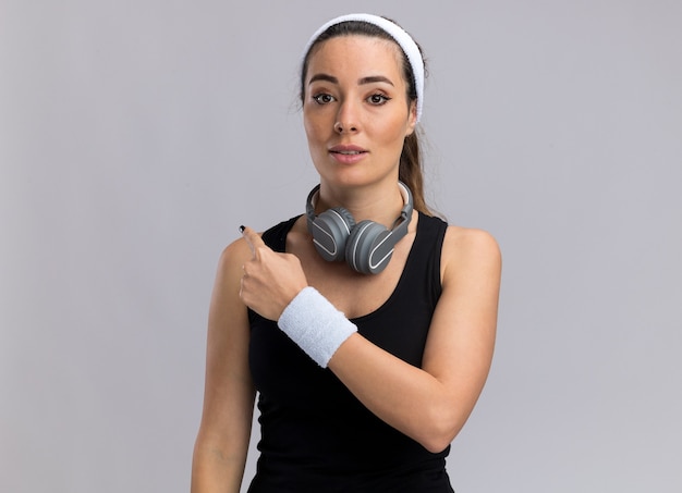 Photo gratuite impressionné jeune fille assez sportive portant un bandeau et des bracelets avec des écouteurs autour du cou pointant derrière