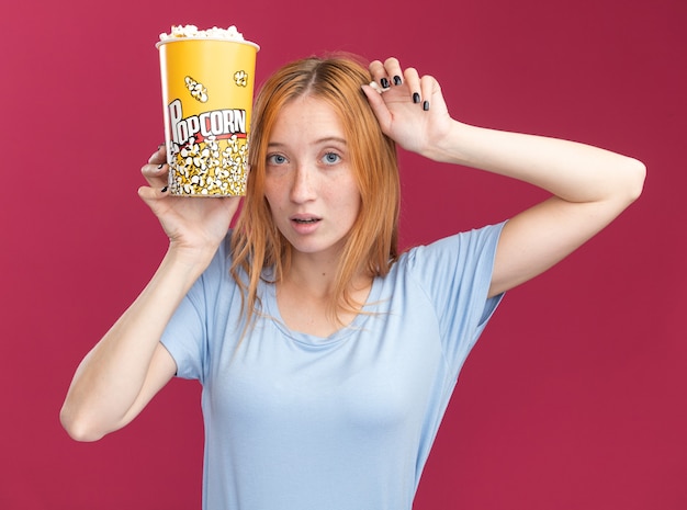 Impressionné jeune fille au gingembre rousse avec des taches de rousseur tenant un seau de pop-corn près du visage sur rose