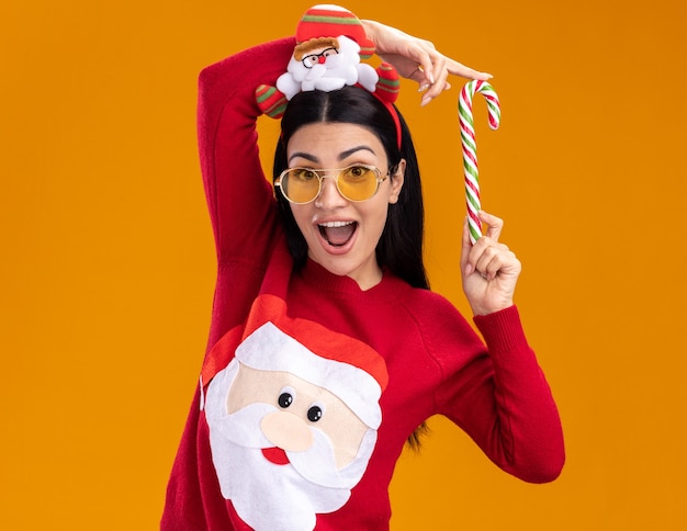 Impressionné jeune fille caucasienne portant bandeau et pull du père Noël avec des lunettes tenant la canne à sucre de Noël traditionnel près de la tête isolée sur le mur orange