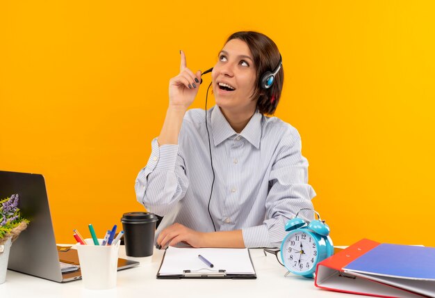 Impressionné jeune fille de centre d'appels portant un casque assis au bureau à la recherche et pointant vers le haut isolé sur orange