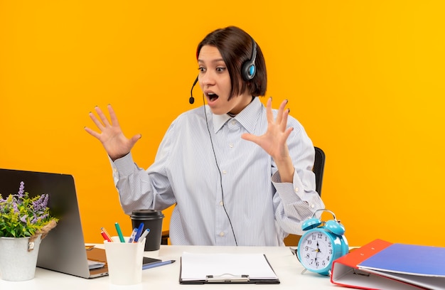 Impressionné Jeune Fille De Centre D'appels Portant Un Casque Assis Au Bureau En Regardant Un Ordinateur Portable Avec Les Mains Surélevées Isolé Sur Orange