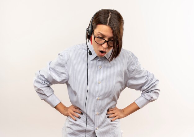 Impressionné jeune fille de centre d'appels portant des lunettes et un casque mettant les mains sur la taille regardant vers le bas isolé sur blanc avec copie espace