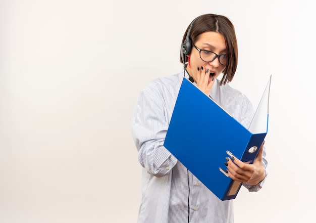 Impressionné jeune fille de centre d'appels portant des lunettes et un casque tenant et regardant le dossier mettant la main sur la bouche isolé sur blanc avec copie