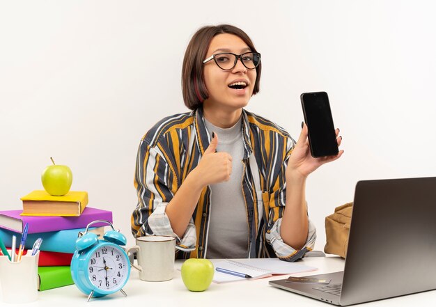 Impressionné jeune fille étudiante portant des lunettes assis au bureau montrant un téléphone mobile et le pouce vers le haut isolé sur blanc