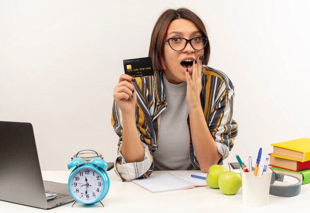 Impressionné jeune fille étudiante portant des lunettes assis au bureau tenant une carte de crédit et mettant la main sur le menton isolé sur blanc