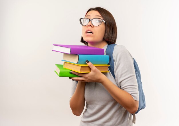 Impressionné jeune fille étudiante portant des lunettes et sac à dos tenant des livres isolés sur blanc