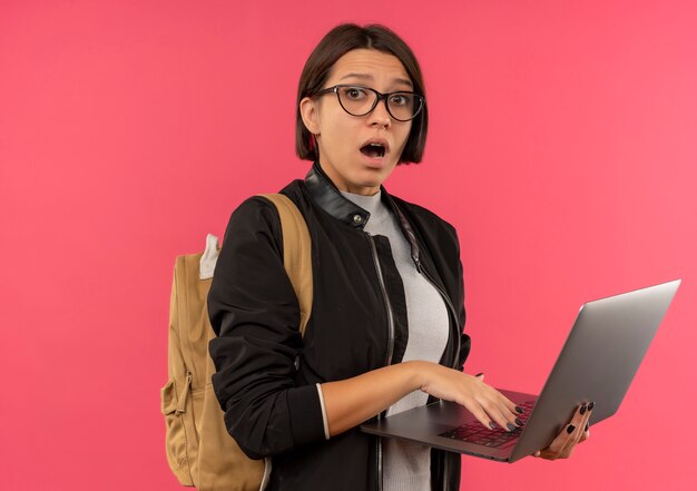 Impressionné jeune fille étudiante portant des lunettes et sac à dos tenant un ordinateur portable isolé sur rose