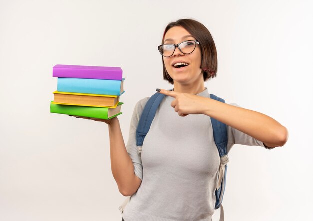 Impressionné jeune fille étudiante portant des lunettes et sac à dos tenant et pointant sur des livres isolés sur blanc