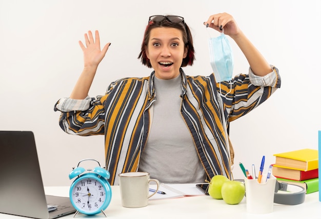 Photo gratuite impressionné jeune fille étudiante portant des lunettes sur la tête assis au bureau tenant un masque et en agitant isolé sur blanc