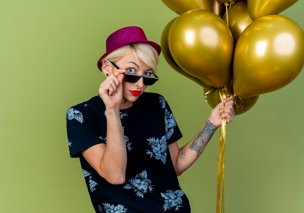 Impressionné jeune fille de fête blonde portant chapeau de fête et lunettes de soleil tenant des ballons et des lunettes en regardant la caméra isolée sur fond vert olive avec espace copie