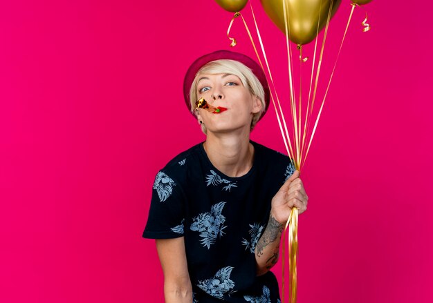 Impressionné jeune fille de fête blonde portant chapeau de fête tenant des ballons et souffleur de fête dans la bouche en regardant la caméra soufflant partie souffleur isolé sur fond cramoisi avec espace de copie