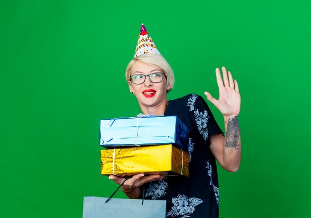 Impressionné jeune fille de fête blonde portant des lunettes et une casquette d'anniversaire regardant côté tenant un sac en papier et des coffrets cadeaux souriant et ne faisant aucun geste isolé sur fond vert avec espace de copie