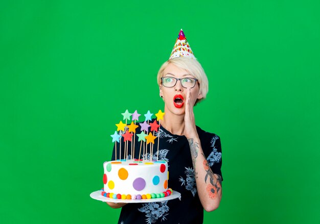 Impressionné jeune fille de fête blonde portant des lunettes et une casquette d'anniversaire tenant un gâteau d'anniversaire avec des étoiles à la recherche de côté chuchotant isolé sur fond vert avec espace copie