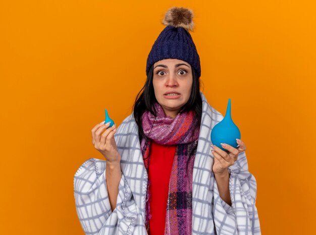 Impressionné jeune fille malade de race blanche portant un chapeau d'hiver et une écharpe enveloppée dans un plaid tenant des lavements isolés sur un mur orange avec copie espace