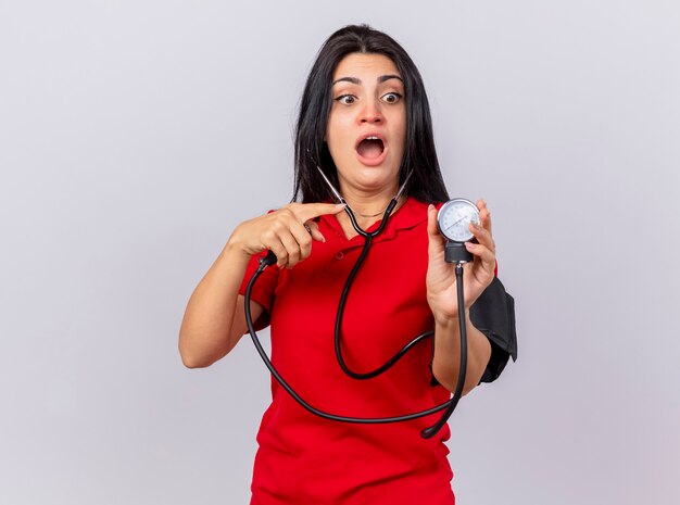 Impressionné jeune fille malade de race blanche portant un stéthoscope mesurant sa pression avec un sphygmomanomètre à la recherche et en le pointant isolé sur fond blanc avec espace de copie