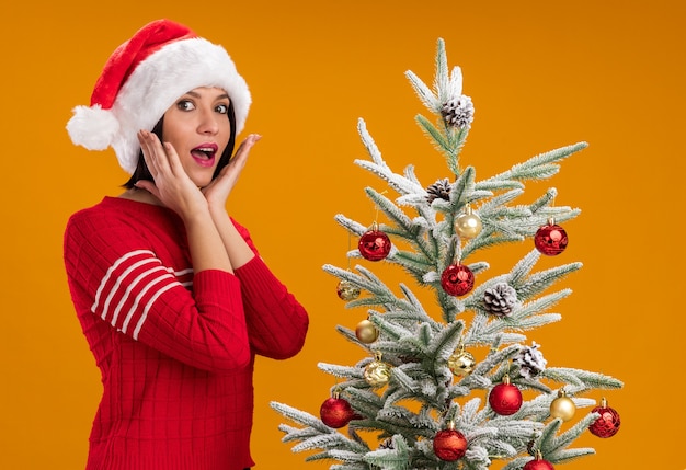 Impressionné jeune fille portant bonnet de Noel debout en vue de profil près de sapin de Noël décoré en gardant les mains près du visage isolé sur mur orange