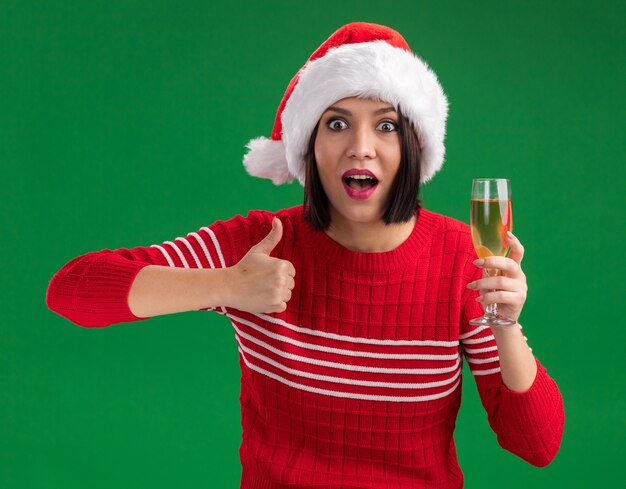 Impressionné jeune fille portant bonnet de Noel tenant un verre de champagne montrant le pouce vers le haut isolé sur le mur vert