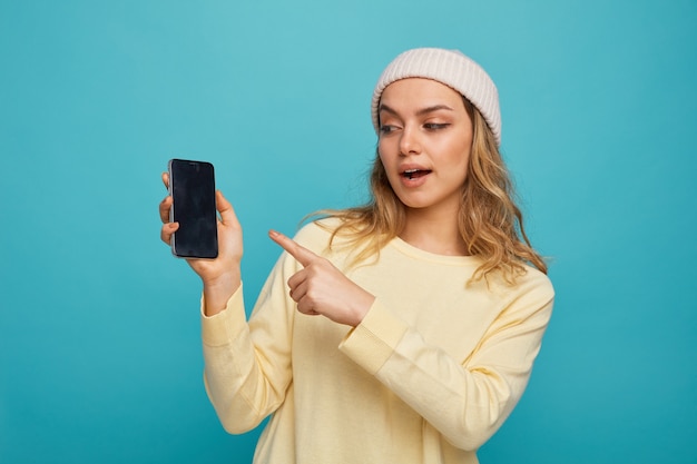 Impressionné jeune fille portant un chapeau d'hiver tenant à la recherche et en pointant sur téléphone mobile