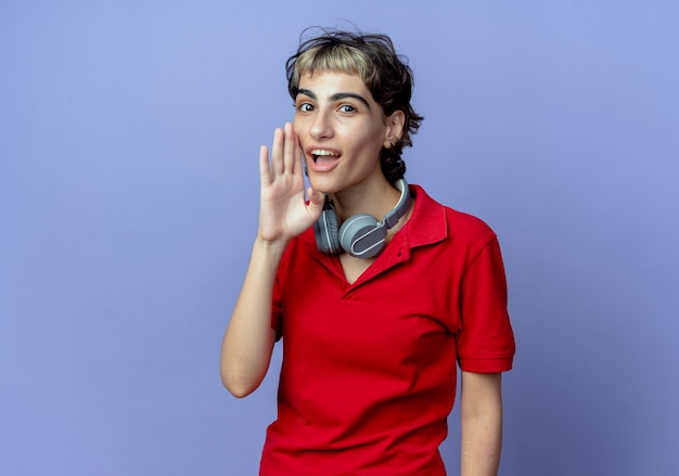 Impressionné jeune fille de race blanche avec coupe de cheveux de lutin portant des écouteurs sur le cou mettant la main près de la bouche chuchotant à la caméra isolée sur fond violet avec espace de copie