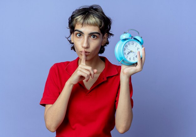 Impressionné jeune fille de race blanche avec coupe de cheveux de lutin tenant un réveil gestes silence isolé sur fond violet avec copie espace