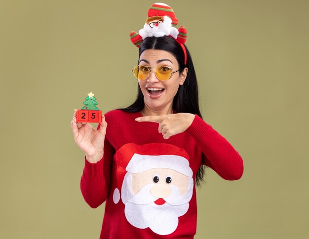 Impressionné jeune fille de race blanche portant bandeau et pull du père Noël avec des lunettes tenant jouet arbre de Noël avec date pointant sur il isolé sur mur vert olive