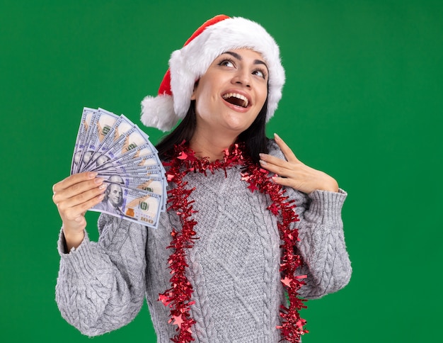 Impressionné jeune fille de race blanche portant chapeau de Noël et guirlande de guirlandes autour du cou tenant de l'argent touchant l'épaule en levant isolé sur fond vert