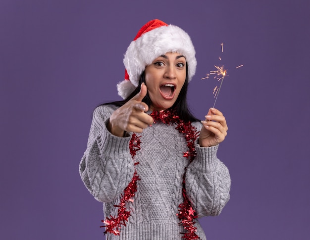 Impressionné jeune fille de race blanche portant chapeau de Noël et guirlande de guirlandes autour du cou tenant un cierge magique isolé sur mur violet avec espace copie