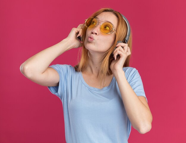 Impressionné jeune fille rousse au gingembre avec des taches de rousseur dans des lunettes de soleil et sur des écouteurs regardant de côté