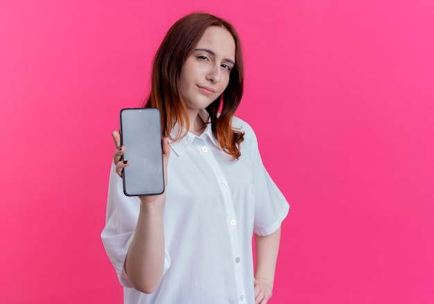 Impressionné jeune fille rousse tenant le téléphone et mettant la main sur la hanche isolé sur fond rose