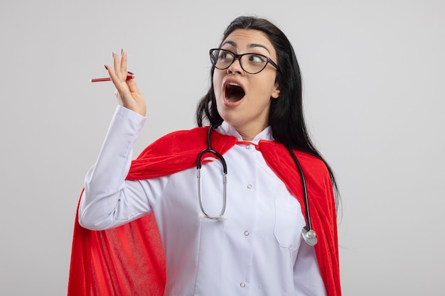 Photo gratuite impressionné jeune fille de super-héros caucasien portant des lunettes et un stéthoscope tenant un crayon rouge à côté isolé sur fond blanc avec espace de copie