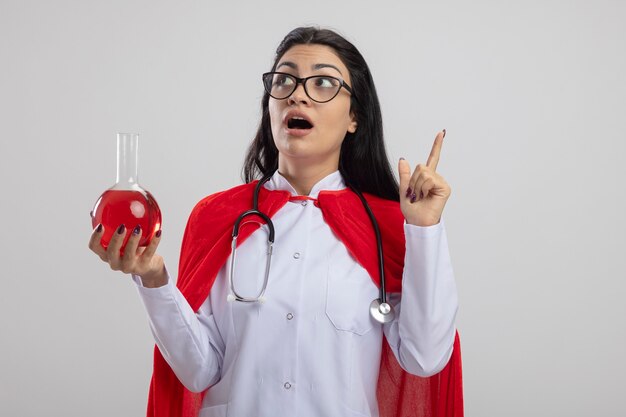 Impressionné jeune fille de super-héros caucasien portant des lunettes et un stéthoscope tenant une fiole chimique avec un liquide rouge à la recherche et pointant vers le haut isolé sur fond blanc avec espace de copie