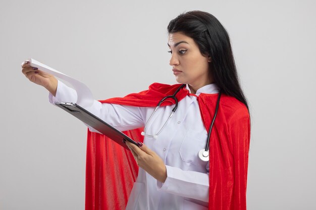 Impressionné jeune fille de super-héros caucasien portant stéthoscope tenant et regardant le presse-papiers isolé sur fond blanc