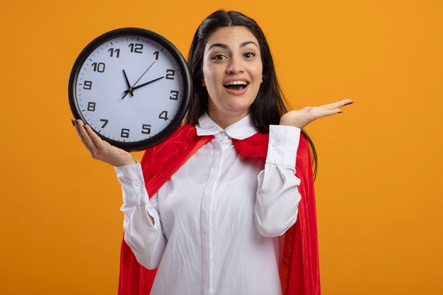 Impressionné jeune fille de super-héros caucasien tenant horloge regardant la caméra montrant la main vide isolée sur fond orange avec espace de copie