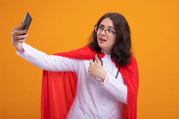 Impressionné jeune fille super-héros caucasienne en cape rouge portant un uniforme de médecin et un stéthoscope avec des lunettes prenant selfie montrant le pouce vers le haut