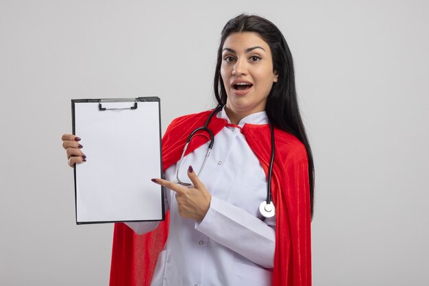 Impressionné jeune fille de super-héros de race blanche portant un stéthoscope montrant le presse-papiers en regardant la caméra isolée sur fond blanc avec copie espace