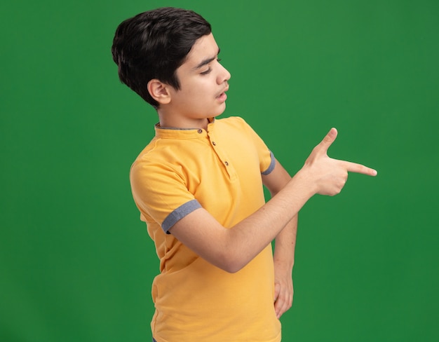 Impressionné jeune garçon caucasien gardant la main sur la taille à la recherche et en pointant sur le côté isolé sur mur vert