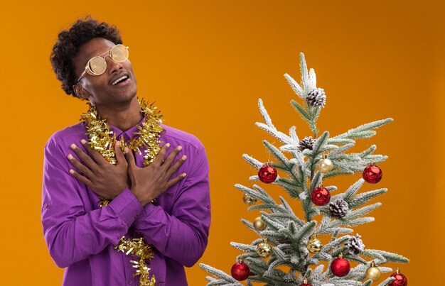 Impressionné jeune homme afro-américain portant des lunettes avec guirlande de guirlandes autour du cou debout près de l'arbre de Noël décoré sur fond orange