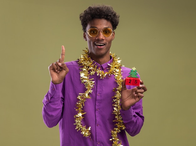 Impressionné jeune homme afro-américain portant des lunettes avec guirlande de guirlandes autour du cou tenant jouet arbre de Noël avec date regardant la caméra pointant vers le haut isolé sur fond vert olive