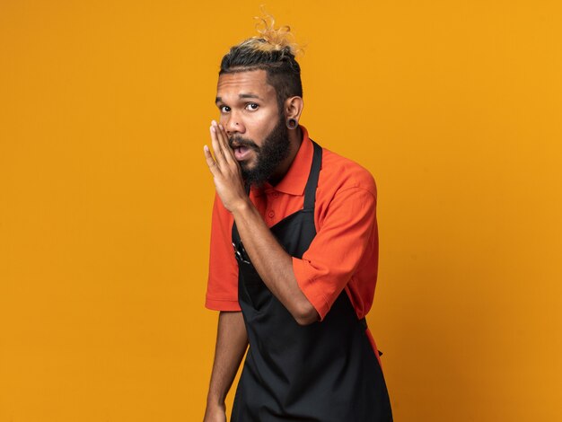 Impressionné jeune homme barbier en uniforme debout dans la vue de profil regardant le chuchotement avant isolé sur mur orange avec espace de copie