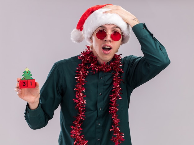 Impressionné Jeune Homme Blond Portant Bonnet De Noel Et Lunettes Avec Guirlande De Guirlandes Autour Du Cou Tenant Un Jouet D'arbre De Noël Avec Date Regardant La Caméra En Gardant La Main Sur La Tête Isolé Sur Fond Blanc