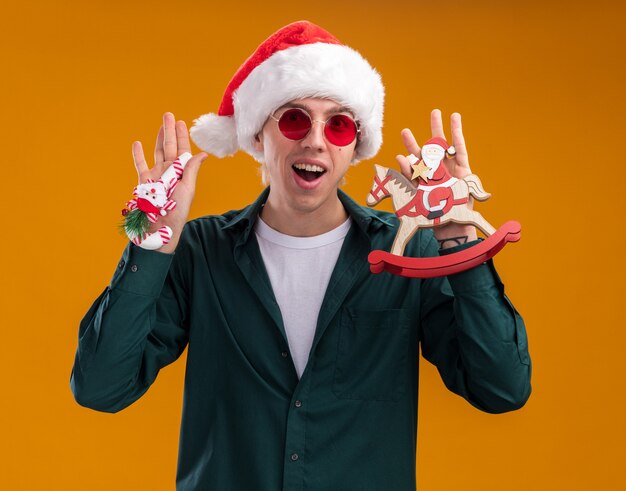 Impressionné jeune homme blond portant Bonnet de Noel et lunettes tenant le Père Noël sur figurine cheval à bascule et ornement de canne à sucre regardant la caméra isolée sur fond orange
