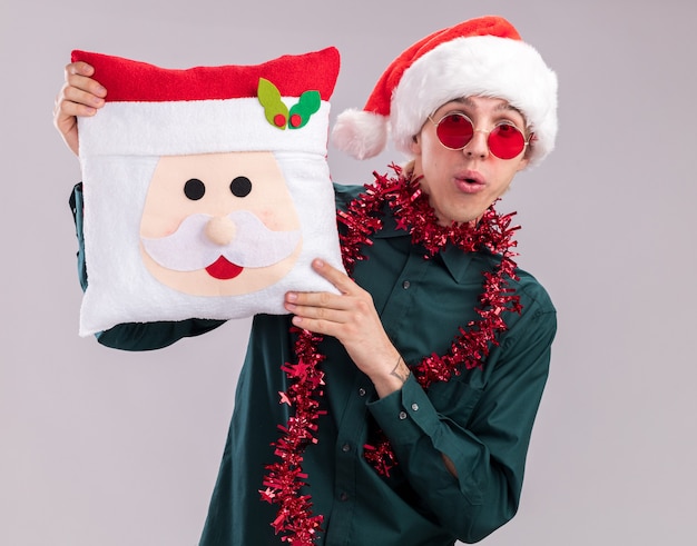 Impressionné jeune homme blond portant un chapeau de père Noël et des lunettes avec une guirlande de guirlandes autour du cou tenant un oreiller de père Noël regardant la caméra isolée sur fond blanc