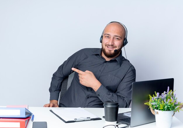 Impressionné jeune homme de centre d'appels chauve portant un casque assis au bureau avec des outils de travail pointant sur le côté isolé sur blanc