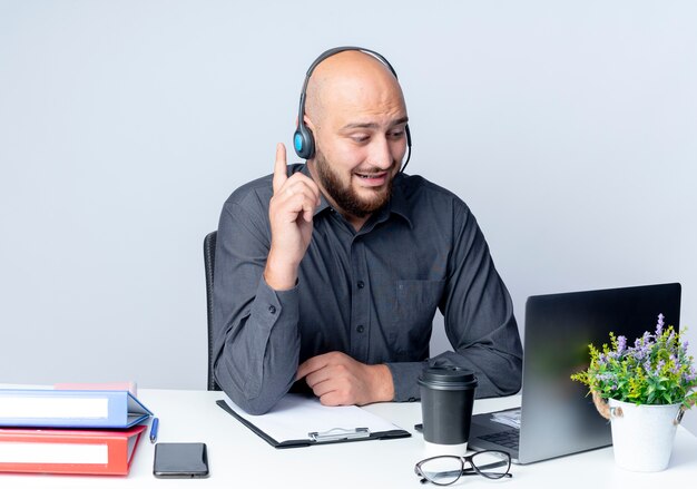 Impressionné jeune homme de centre d'appels chauve portant un casque assis au bureau avec des outils de travail en regardant ordinateur portable et en levant le doigt isolé sur blanc