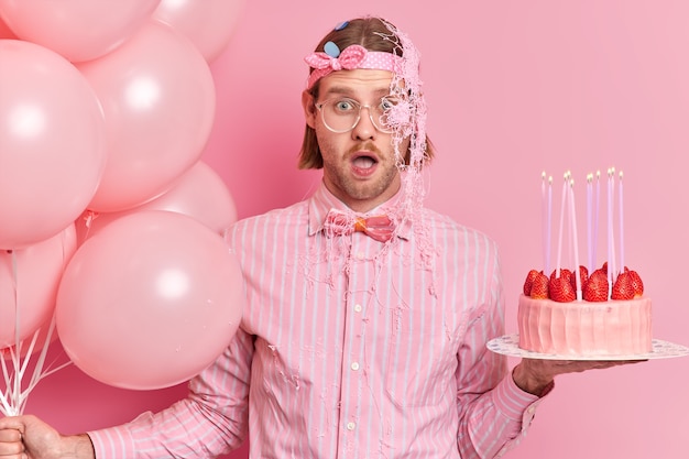 Impressionné, un jeune homme choqué dans des vêtements de fête regarde les yeux obstrués garde la bouche ouverte reçoit les félicitations inattendues d'un ami enduit de crème tient un gâteau d'anniversaire et des ballons gonflés