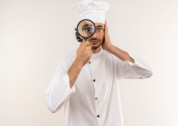 Impressionné jeune homme cuisinier portant l'uniforme de chef et des lunettes avec loupe