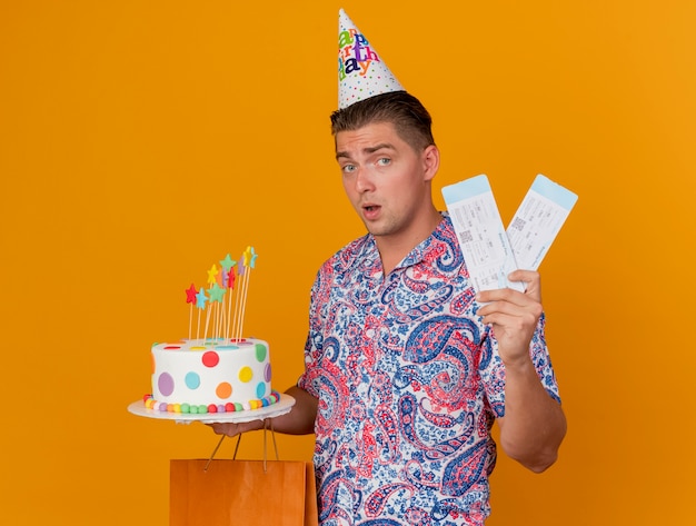 Impressionné jeune homme de fête portant une casquette d'anniversaire tenant un sac-cadeau avec un gâteau et des billets isolés sur orange